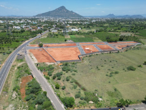 An aerial view of a large plot of red soil near Tiruvannamalai Main Road, divided into segments for potential residential development. The surrounding landscape features green fields, paved roads, and a distant hill under a clear sky. The layout suggests structured urban expansion amidst natural surroundings, ideal for showcasing real estate growth and investment potential.