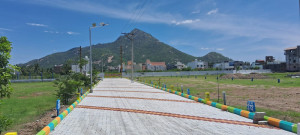 A paved walkway bordered with colorful edging leads into a residential area featuring a mix of houses and buildings. The path is lined with streetlights and small plants, while a lush green mountain rises in the background beneath a clear blue sky. Plot markers labeled 272 and 293 are visible beside the path.